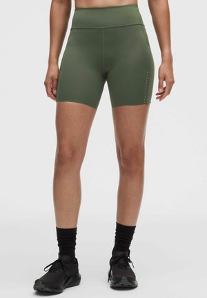 Donna che indossa pantaloncini sportivi a vita alta color verde oliva, calzini neri e scarpe da corsa nere, in piedi davanti a uno sfondo chiaro e uniforme.