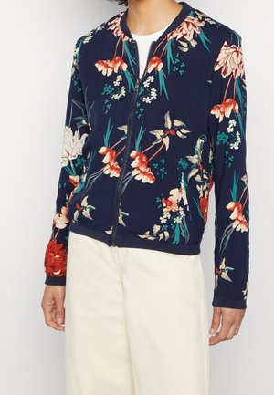 Person trägt dunkelblaue Jacke mit Reißverschluss, rotem, weißem und grünem Blumenmuster und cremefarbenen Hosen vor weißem Hintergrund.