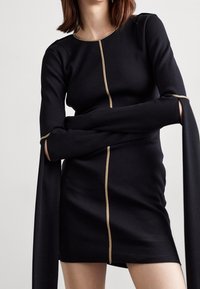 Svart ribbade klänning med långa ärmar och slitsar, framhävd av guldtrim längs den främre sömmen. Tätt sittande silhuett, mjuk textur.