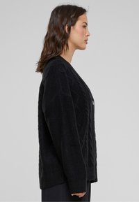 Zwarte gebreide cardigan met kabelpatroon, ronde halslijn, knoopsluiting en geribde manchetten. Losse pasvorm met een zachte textuur.