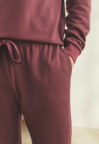 Burgundi színű, waffle textúrájú pulóver és hozzá illő nadrág állítható derékkal. Kényelemre tervezve, bordázott mandzsettákkal és laza szabással.