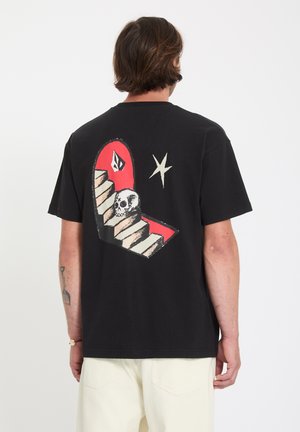 Homme portant un t-shirt noir avec un motif d'escaliers, de crâne, d'étoile et de forme géométrique dans le dos, associé à un pantalon de couleur claire.