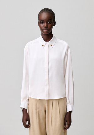 Mujer con el pelo trenzado que lleva una camisa blanca abotonada con botones dorados en el cuello y pantalones plisados beige, de pie frente a un fondo liso.