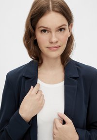 Navy blazer with a lapel design, lightweight fabric, paired with a white ribbed shirt; the model gently holds the blazer's collar.