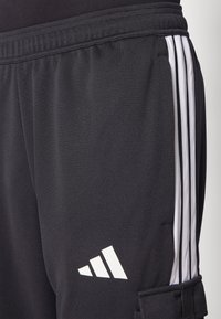 Melni sporta bikses, kas izgatavotas no teksturēta auduma, ar baltu trīs svītru dizainu un logotipu kreisajā pusē. Jostasvieta ietver elastīgu detalizāciju.