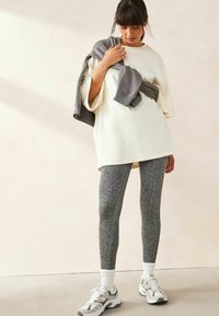 Přes oversized krémová mikina s širokými rukávy, kombinovaná s texturovanými šedými legínami a bílými sportovními ponožkami, doplněná šedými teniskami.
