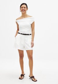 White off-the-shoulder fitted top with gathered texture, paired with white tailored shorts featuring pleats, and a black belt. Brown heeled sandals.