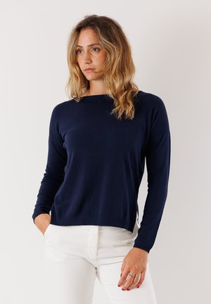 Donna con capelli biondi ondulati che indossa un maglione a maniche lunghe blu navy e pantaloni bianchi, in piedi con una mano in tasca su uno sfondo bianco.
