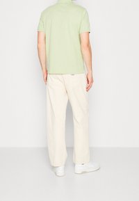 Un homme portant une chemise à manches courtes vert clair, un pantalon ample écru et des baskets blanches se tient de dos contre un fond uni.