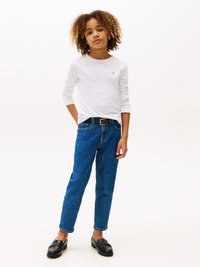 Langermet hvit skjorte, blå teksturerte jeans, svart belte og svarte mokasiner. Barnet poserer med hendene i lommene, og viser frem et avslappet antrekk.