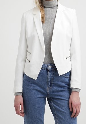 Kobieta w białej, dopasowanej marynarce z bocznymi zamkami, na szarym golfie i niebieskich, szerokich dżinsach z wysokim stanem na jednolitym tle.