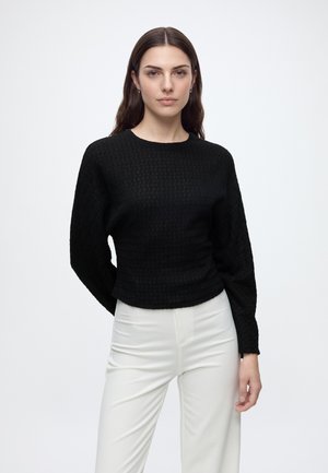 Jeune femme aux longs cheveux bruns, portant un haut noir texturé à manches longues et un pantalon blanc, debout devant un fond uni.