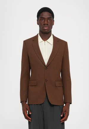 Mann in braunem Blazer über einem cremefarbenen Reißverschlusshemd und dunklen grauen Hosen, steht vor einem schlichten weißen Hintergrund.