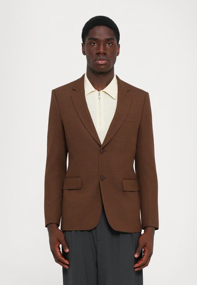 Homme portant un blazer marron sur une chemise zippée crème et un pantalon gris foncé, se tenant contre un fond blanc uni.