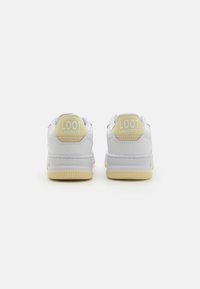 Hvite Lacoste-sneakers med gule hælakkenter, med en glatt skinntekstur, avrundet form og subtil branding på baksiden.
