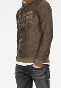 Olivově zelená mikina s texturovanou látkou, šňůrkami a decentním logem. Kombinována s tmavě šedými džíny se vzdušným střihem.