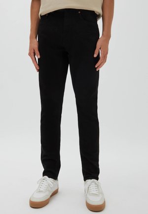 Mann trägt schwarze Slim-Fit-Jeans und weiße Turnschuhe, steht vor einem schlichten hellen Hintergrund.