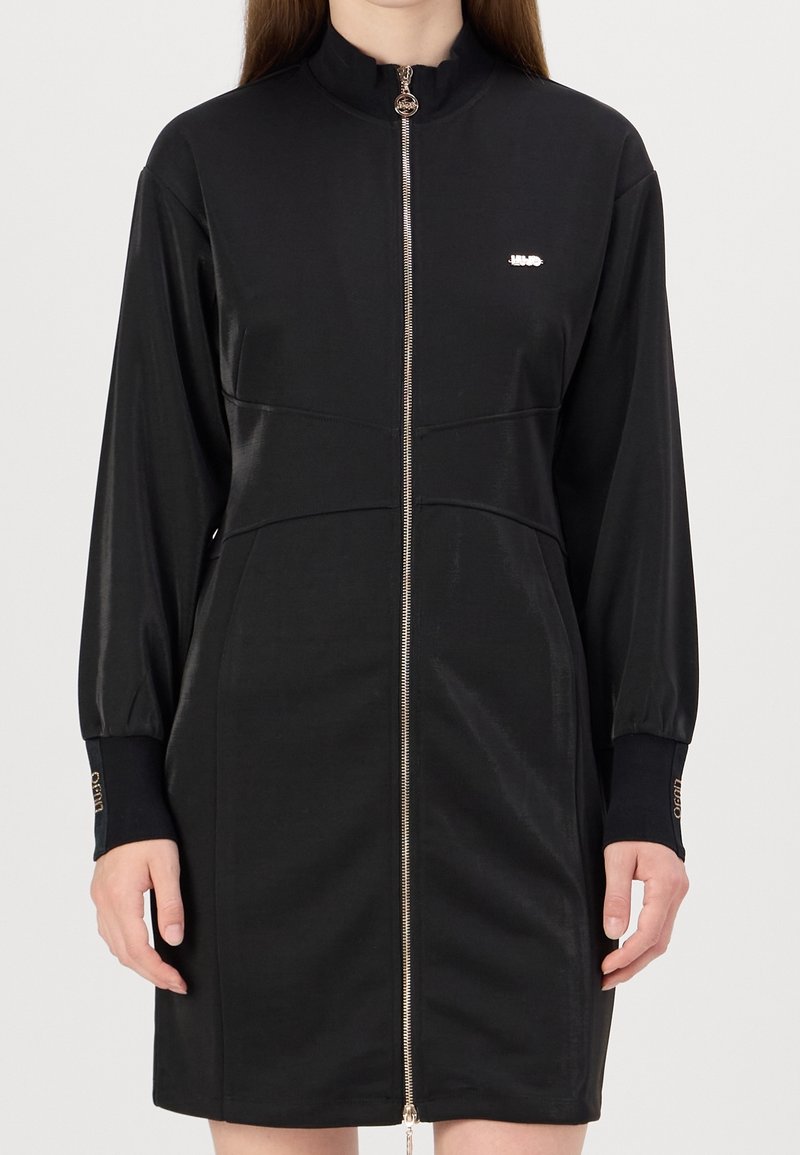 Zwarte ritsjurk met een hoge kraag, lange mouwen en een metallic logo-accent. Soepele stof met een getailleerde pasvorm en zijzakken.
