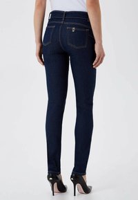 Jean skinny en denim bleu foncé, avec une taille mi-haute, un design à cinq poches et des coutures jaunes discrètes. Porté avec des talons hauts noirs.