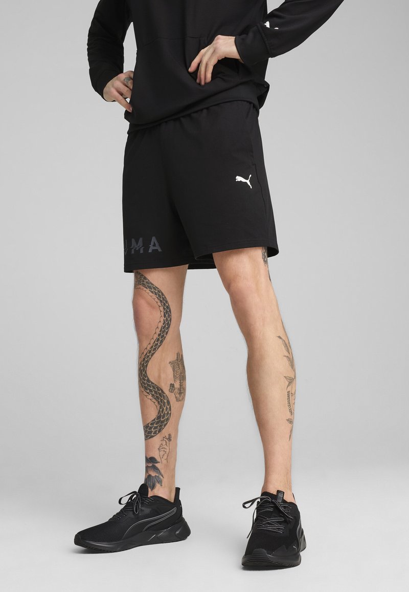 Shorts de sport negre cu talie elastică, având un logo Puma în litere albe și gri, asociați cu pantofi sport negri.