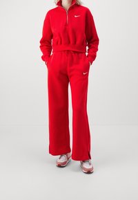 Röd Nike-träningsdräkt bestående av en zip-up-overall med hög krage och löst sittande byxor. Ställd mot en neutral bakgrund. Accentuerad med vit logotyp.