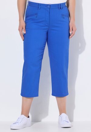 Blaue kurze Hosen mit vorderen Reißverschlusstaschen, getragen mit weißen Turnschuhen und einem weißen Oberteil, stehend vor einem schlichten hellen Hintergrund.