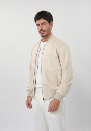 Férfi bézs zipzáras bomber dzsekiben, fehér inggel és fehér nadrággal, világos egyszínű háttér előtt állva.