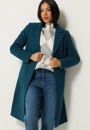 Femme portant un manteau long bleu sarcelle par-dessus un pull blanc et gris à motifs avec un jean bleu, debout, les mains tenant les revers du manteau.