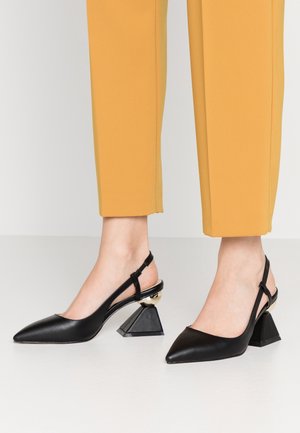 Jambes portant un pantalon moutarde et des escarpins slingback noirs à bout pointu avec talons blocs géométriques et accents métalliques.