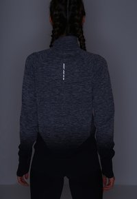 Vrouw met gevlochten haar, gekleed in een donkergrijze sportjack met lange mouwen en duimgaten, van achteren gezien tegen een effen donkere achtergrond.