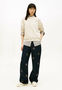 Felpa cremosa, camicia a quadri sotto, jeans a gamba larga blu scuro con emblemi. Sneakers bianche completano l'outfit casual.