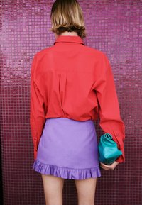 Persona con cabello rubio corto de pie frente a una pared de azulejos morados, lleva una camisa roja, falda morada con dobladillo con volantes y sostiene un bolso de mano azul verdoso.