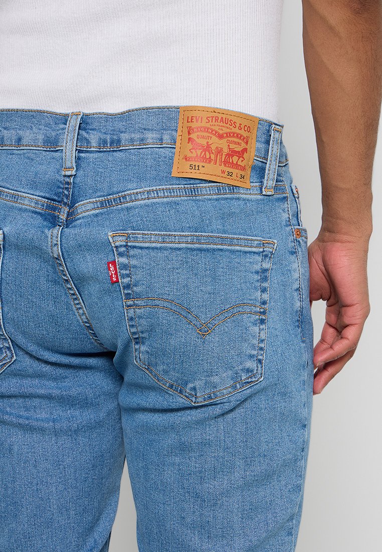 Jean en denim bleu clair à coupe slim, avec une étiquette en cuir marron à l'arrière comportant des graphismes rouges et orange, et des poches arrière standard.