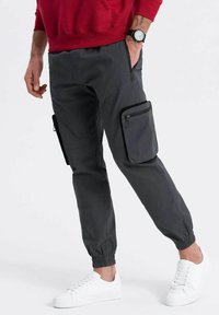 Cargo-Hosen in Dunkelgrau, mit elastischen Bündchen, mehreren Reißverschlusstaschen und einer glatten Textur. Kombiniert mit weißen Sneakers.