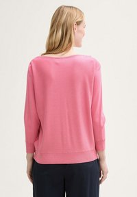 Pulover tricotat roz cu mâneci trei sferturi, guler larg și tiv ribcat. Textură netedă, asortat cu pantaloni negri.