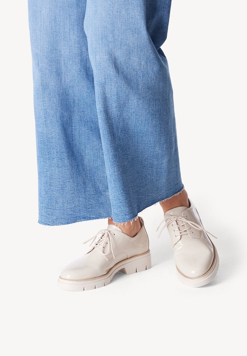 Chaussures en cuir crème à lacets avec une finition brillante, une semelle épaisse texturée et un design minimaliste, portées avec un pantalon large en denim bleu.