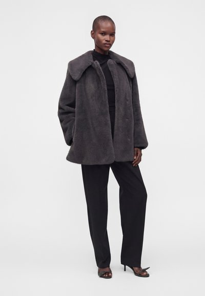 Femme portant un manteau oversize en fausse fourrure gris foncé, un col roulé noir, un pantalon noir et des talons ouverts noirs, debout devant un fond clair uni.