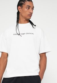 T-shirt branco de manga curta feito de algodão com decote redondo. Apresenta o texto "SYSTEM TEMP. CRITICAL" em cinza escuro na parte da frente.