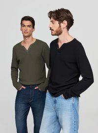 Dwóch mężczyzn nosi koszule henley z długim rękawem: jedna oliwkowa z guzikami, druga czarna. Do nich dopasowane są niebieskie jeansy.