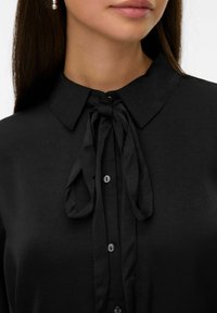 Zwarte blouse met een kraag en een striksluiting bij de hals, met een gladde textuur en knopen langs de voorkant.