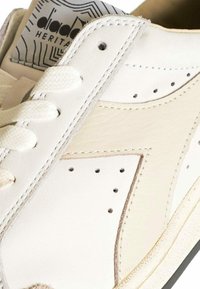 Sneaker bianca in pelle con una texture liscia, dotata di perforazioni, lacci piatti e suola scura in contrasto. Il logo sulla linguetta è visibile.