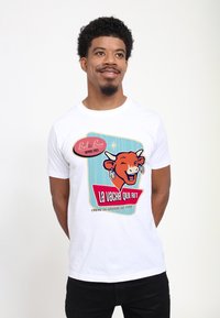 Una maglietta bianca presenta un graphic colorato con un logo di una mucca sorridente, uno sfondo blu e la scritta "La Vache Qui Rit" in grassetto rosso e blu.