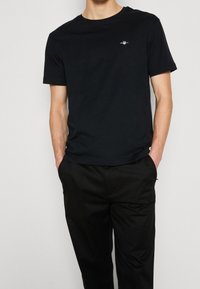 Maglietta nera in cotone con maniche corte, scollo rotondo e piccolo logo sul petto, abbinata a pantaloni neri. Tessuto dalla texture liscia.