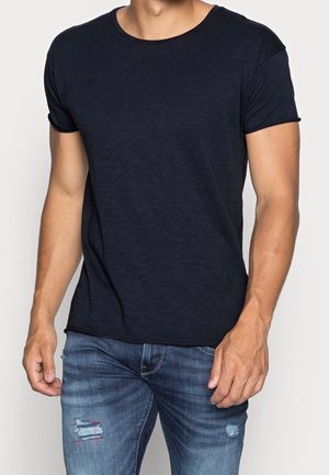 Mand iført en ensfarvet sort t-shirt med korte ærmer og blå slidte jeans mod en lys baggrund.