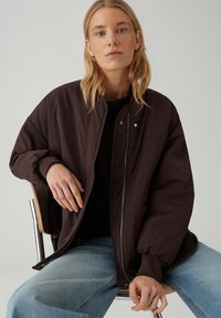 Vrouw met blond haar die een donkerbruine oversized jas en lichtblauwe spijkerbroek draagt, zittend op een houten stoel tegen een effen achtergrond.