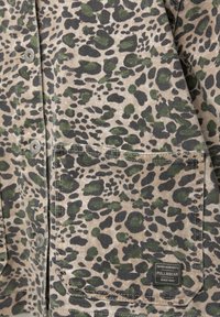 Tissu à motif camouflage beige et vert avec une poche avant et des boutons-pression gris sur un vêtement.