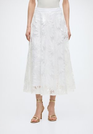 Λευκή midi φούστα με μεγάλο διακοσμητικό δαντελένιο πάνω μέρος με λουλούδια, φορεμένη με χρυσά σανδάλια με ψηλό τακούνι.