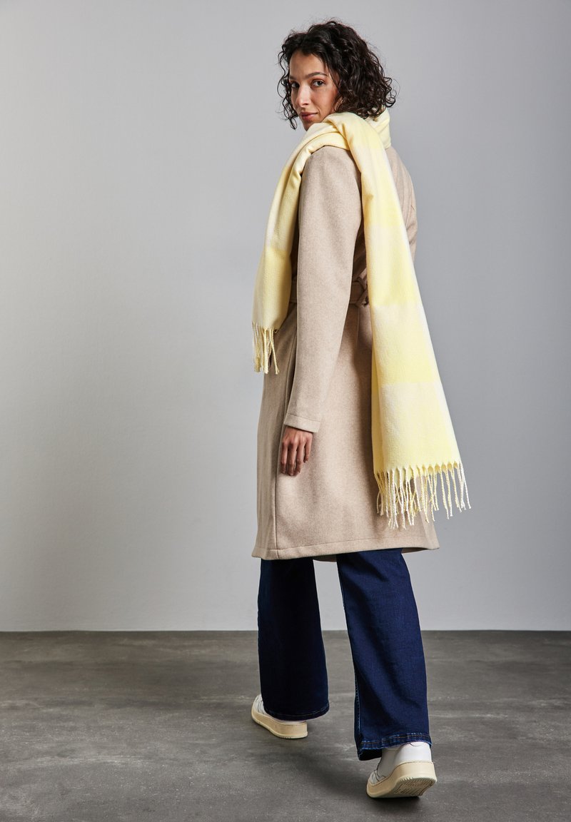 Cappotto beige con taglio su misura, sciarpa gialla a righe drappeggiata su una spalla, jeans a gamba larga blu scuro e sneakers bianche.