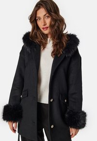 Black wool coat with a faux fur hood and cuffs, featuring silver button hardware and side pockets. Textured knit sweater underneath.