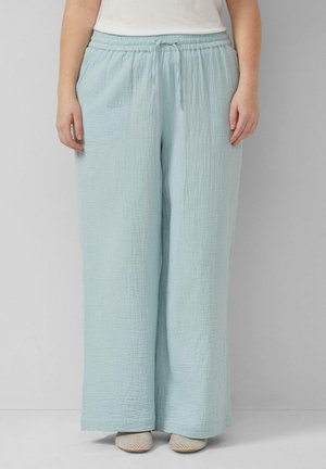 Loszittende lichtblauwe broek met textuur, elastische tailleband en trekkoord, gedragen met witte schoenen en een wit topje, staand tegen een effen muur.